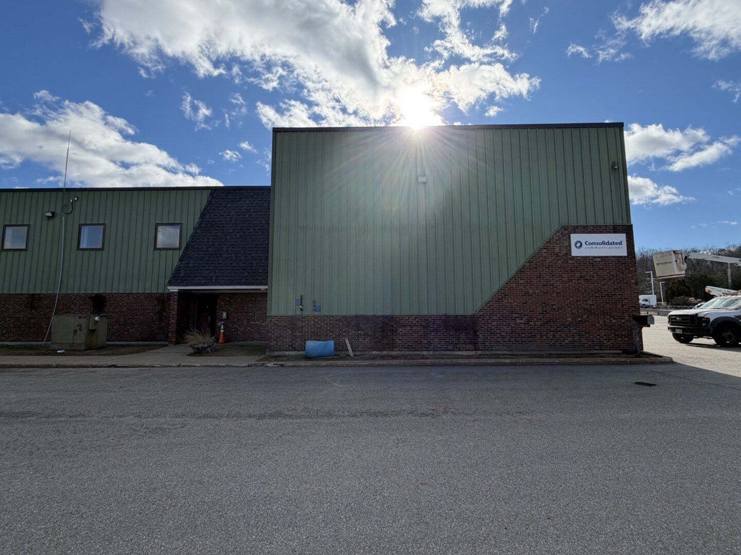 Building with windows and a name board, a car parked in front of it, a group of trees, some poles