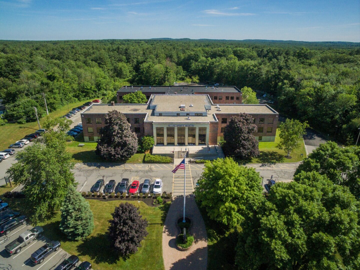 Aerial view of a large building with a flagpole in front of it, surrounded by trees, grass, plants